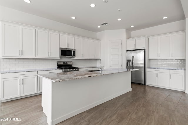 kitchen with tasteful backsplash, light stone counters, appliances with stainless steel finishes, white cabinets, and a kitchen island with sink