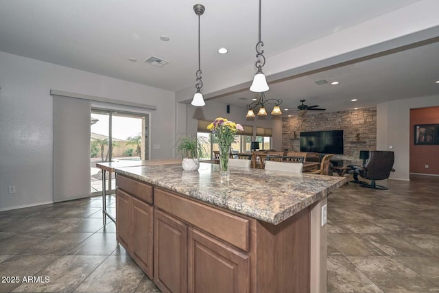 kitchen with a stone fireplace, a center island, pendant lighting, ceiling fan, and light stone countertops