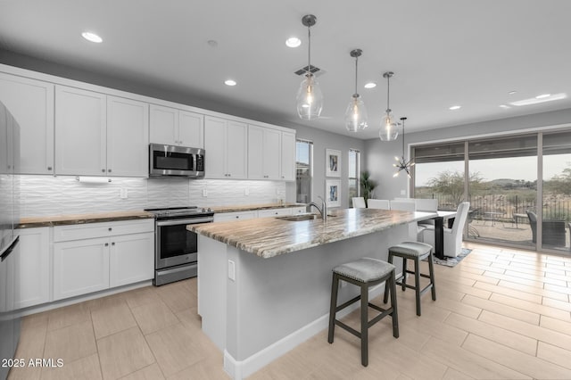 kitchen with an island with sink, white cabinetry, hanging light fixtures, stainless steel appliances, and light stone countertops