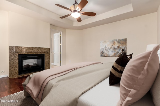 bedroom featuring dark wood finished floors, a multi sided fireplace, a ceiling fan, and a tray ceiling