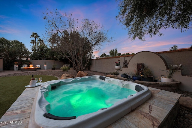 pool at dusk with hot tub deck surround, a fire pit, and a fenced backyard