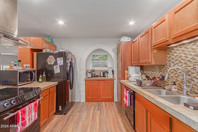 kitchen with wall chimney range hood, decorative backsplash, light wood-type flooring, black appliances, and sink