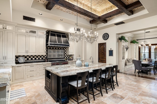 kitchen featuring a center island with sink, wall chimney range hood, hanging light fixtures, tasteful backsplash, and light stone counters