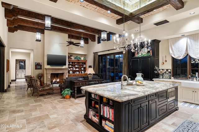 kitchen with beamed ceiling, light stone counters, an island with sink, and a high ceiling