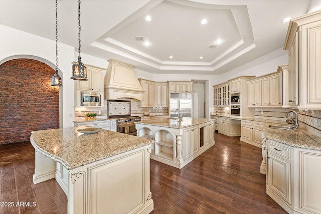 kitchen featuring built in appliances, a raised ceiling, cream cabinets, and custom exhaust hood