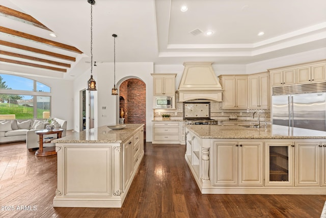 kitchen featuring an island with sink, built in appliances, cream cabinets, custom exhaust hood, and a sink