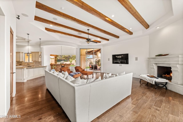 living room with a warm lit fireplace, ceiling fan, arched walkways, beamed ceiling, and dark wood finished floors