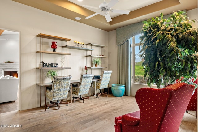 interior space featuring a warm lit fireplace, ceiling fan, a tray ceiling, and wood finished floors