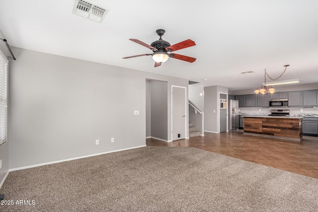 unfurnished living room featuring dark colored carpet, visible vents, stairway, baseboards, and ceiling fan with notable chandelier