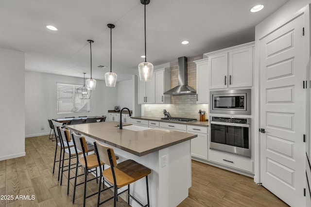 kitchen featuring appliances with stainless steel finishes, a kitchen island with sink, light wood-type flooring, wall chimney range hood, and backsplash