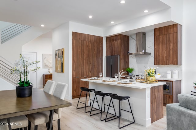 kitchen featuring wall chimney range hood, stainless steel fridge, an island with sink, a breakfast bar, and light wood-type flooring