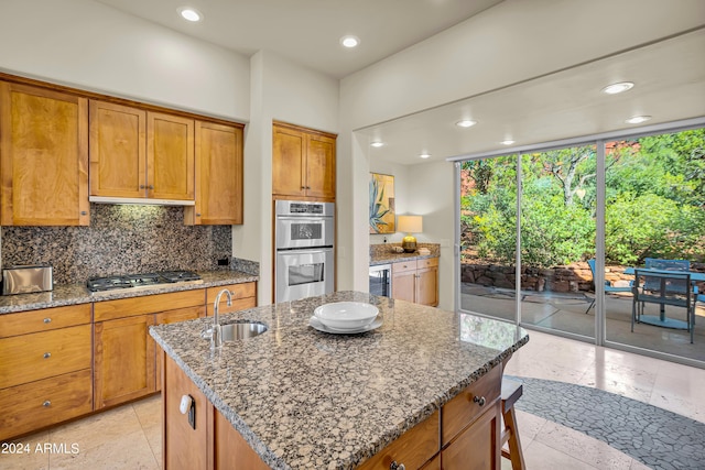 kitchen with a sink, stainless steel appliances, brown cabinetry, a center island with sink, and backsplash