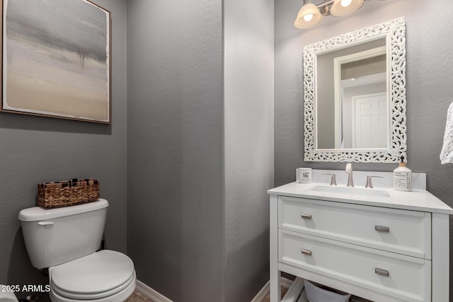 half bathroom featuring baseboards, a textured wall, vanity, and toilet