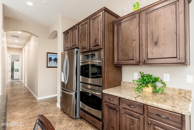 kitchen featuring light stone countertops, dark brown cabinetry, arched walkways, stainless steel appliances, and baseboards