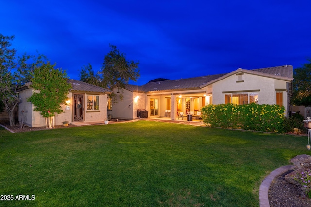 back of house at night with a lawn, a tile roof, a patio area, and stucco siding