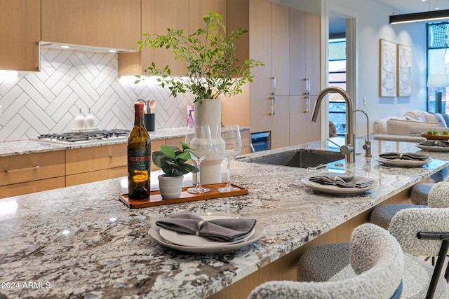 kitchen with backsplash, sink, light stone countertops, light brown cabinetry, and stainless steel gas cooktop