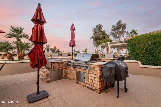 patio terrace at dusk featuring area for grilling, a pergola, and exterior kitchen