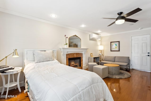 bedroom with crown molding, a wall mounted AC, a tile fireplace, ceiling fan, and hardwood / wood-style floors