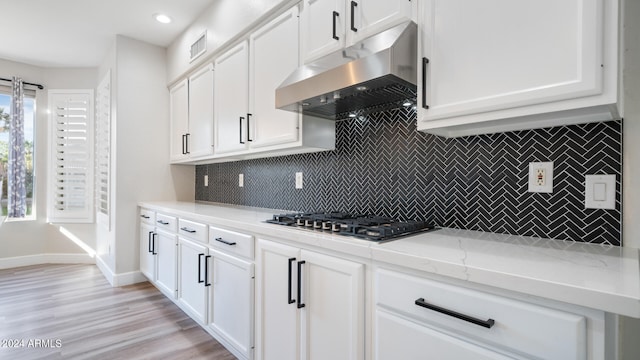 kitchen featuring ventilation hood, stainless steel gas stovetop, white cabinets, decorative backsplash, and light stone counters