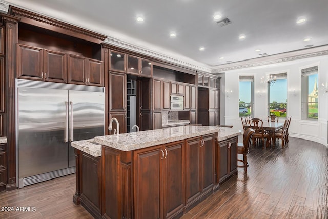 kitchen with dark brown cabinetry, light stone countertops, dark wood-type flooring, stainless steel built in refrigerator, and a center island with sink