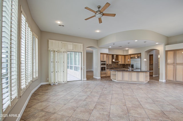 kitchen featuring appliances with stainless steel finishes, backsplash, wall chimney exhaust hood, dark countertops, and a center island with sink