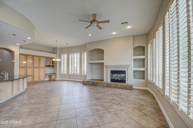 unfurnished living room with ceiling fan with notable chandelier, visible vents, baseboards, built in features, and a glass covered fireplace