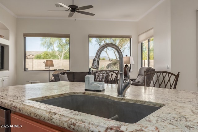 kitchen with light stone counters, a sink, a ceiling fan, open floor plan, and ornamental molding