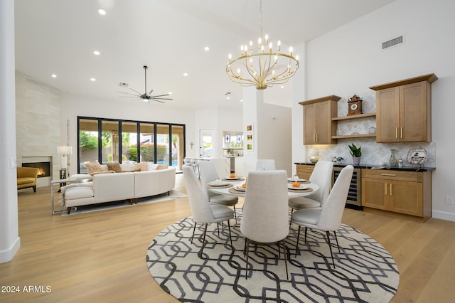 dining area with a large fireplace, beverage cooler, a high ceiling, ceiling fan with notable chandelier, and light wood-type flooring