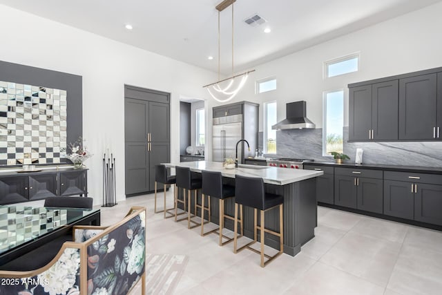 kitchen with built in fridge, pendant lighting, a breakfast bar area, a kitchen island with sink, and wall chimney exhaust hood