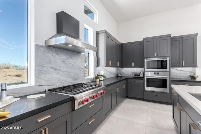 kitchen with wall chimney exhaust hood, tasteful backsplash, light tile patterned floors, dark stone countertops, and appliances with stainless steel finishes