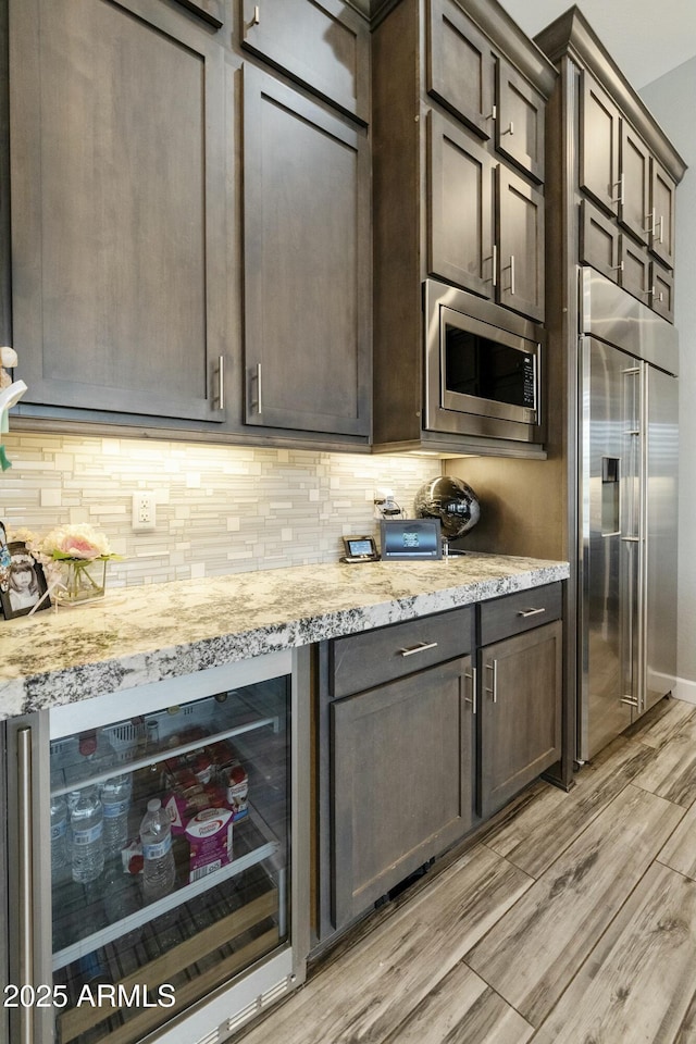 kitchen with tasteful backsplash, beverage cooler, built in appliances, light wood-type flooring, and light stone counters