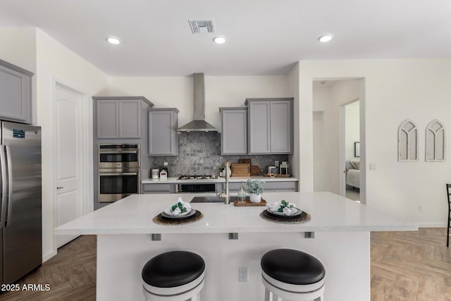 kitchen featuring wall chimney exhaust hood, a center island with sink, parquet floors, appliances with stainless steel finishes, and a kitchen breakfast bar