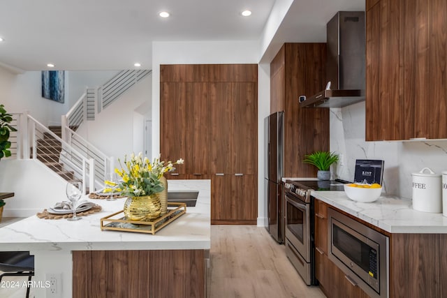 kitchen with light stone countertops, stainless steel appliances, light hardwood / wood-style floors, and wall chimney range hood