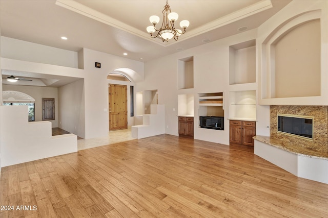 unfurnished living room with ceiling fan with notable chandelier, a raised ceiling, built in shelves, a fireplace, and light hardwood / wood-style floors