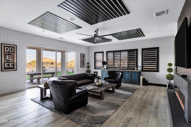 living room with brick wall, hardwood / wood-style flooring, ceiling fan, a glass covered fireplace, and visible vents