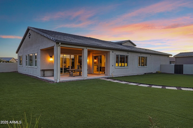 back of house at dusk featuring fence, a lawn, a patio, and stucco siding