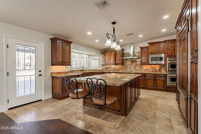 kitchen featuring a kitchen island, appliances with stainless steel finishes, stone countertops, decorative backsplash, and wall chimney range hood