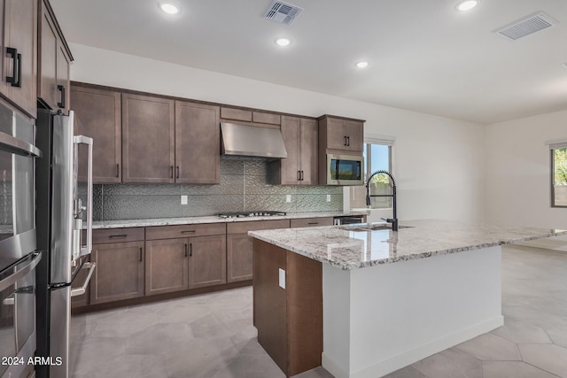 kitchen with decorative backsplash, light stone counters, an island with sink, sink, and stainless steel appliances