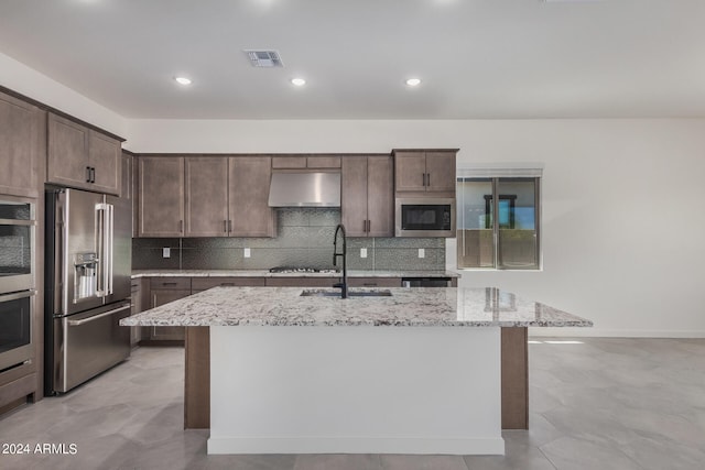 kitchen with sink, an island with sink, stainless steel appliances, light stone counters, and extractor fan