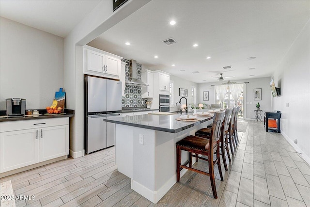 kitchen featuring dark countertops, visible vents, a center island with sink, appliances with stainless steel finishes, and wall chimney exhaust hood