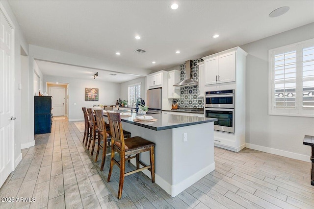 kitchen featuring wood finish floors, a breakfast bar, backsplash, appliances with stainless steel finishes, and wall chimney range hood