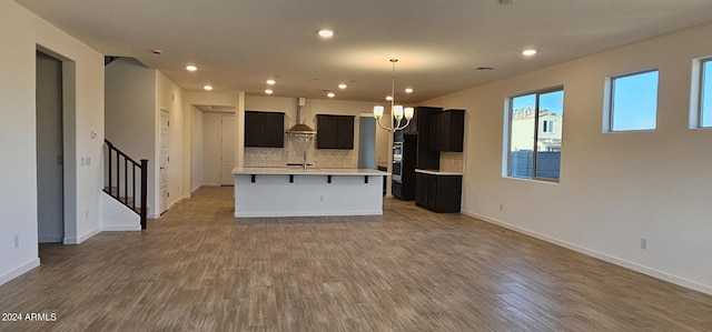 kitchen featuring wall chimney exhaust hood, an inviting chandelier, tasteful backsplash, a center island with sink, and hardwood / wood-style floors
