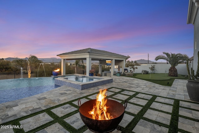 pool at dusk featuring a patio, an in ground hot tub, pool water feature, an outdoor fire pit, and a gazebo