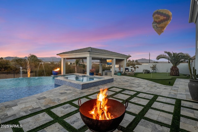 pool at dusk featuring pool water feature, a gazebo, an in ground hot tub, a fire pit, and a patio