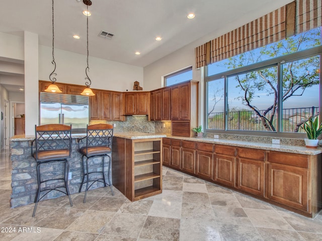 kitchen featuring hanging light fixtures, built in refrigerator, backsplash, light stone countertops, and a high ceiling