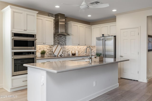 kitchen with wall chimney exhaust hood, sink, light hardwood / wood-style flooring, appliances with stainless steel finishes, and a kitchen island with sink