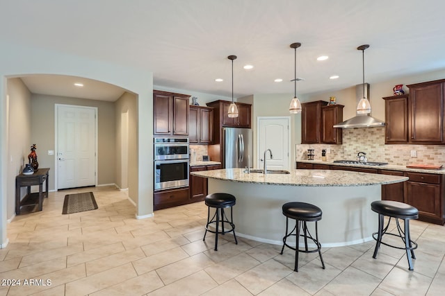 kitchen with pendant lighting, an island with sink, sink, wall chimney range hood, and stainless steel appliances