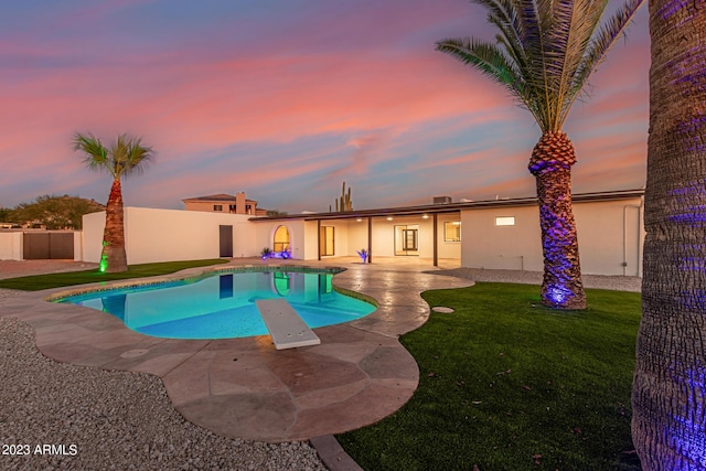 pool at dusk with a patio area, a diving board, and a lawn