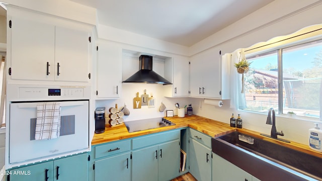 kitchen with butcher block countertops, wall chimney range hood, white cabinetry, and oven