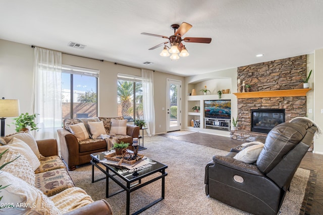 living room with a stone fireplace, a textured ceiling, carpet floors, built in features, and ceiling fan